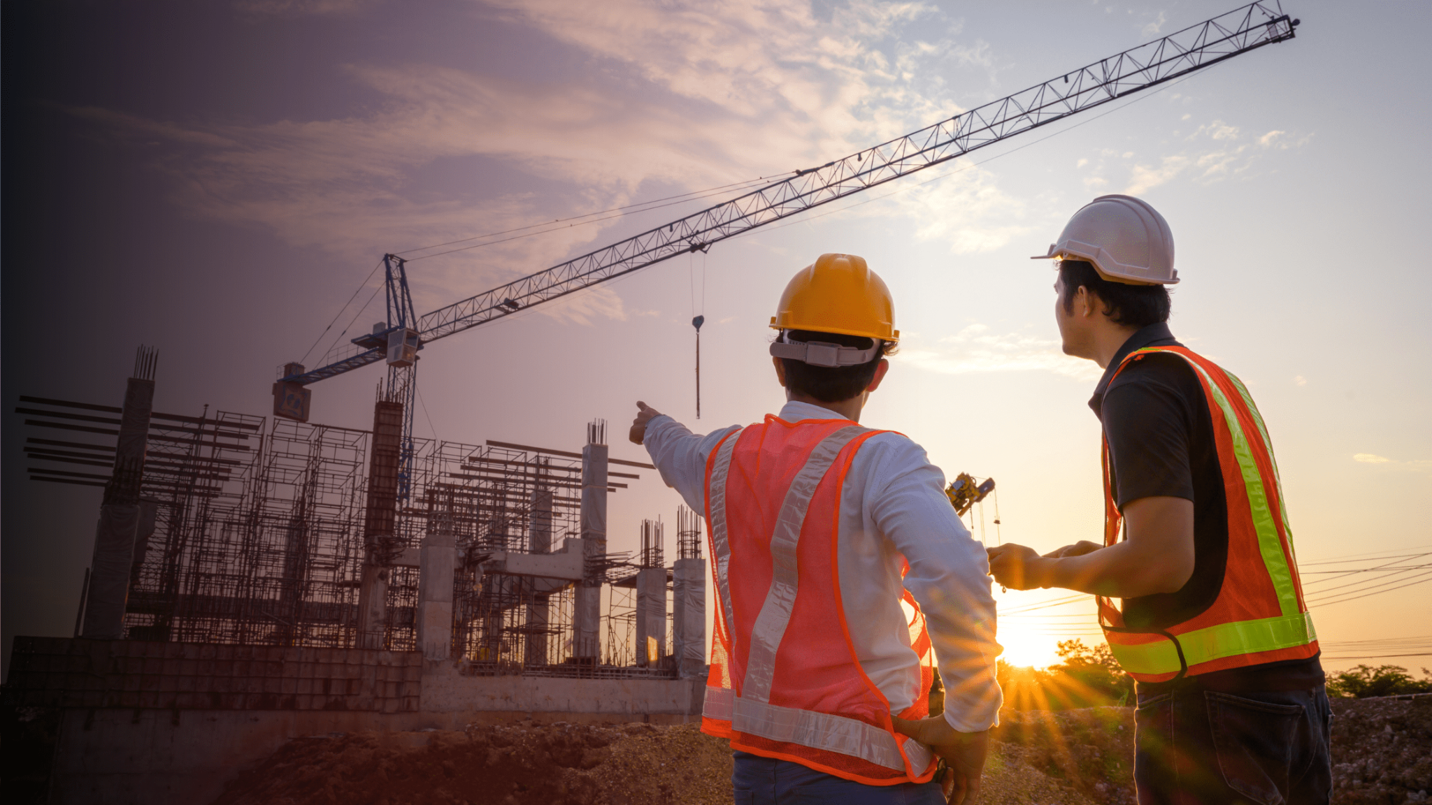 Two construction professionals in hard hats and high-visibility vests discussing works beside a building site and tower crane