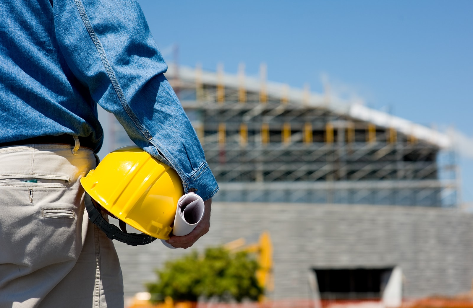 A person holding rolled drawings and a yellow hard hat beside a building under construction