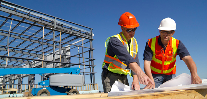 Two construction professionals checking drawings beside a steel-framed building