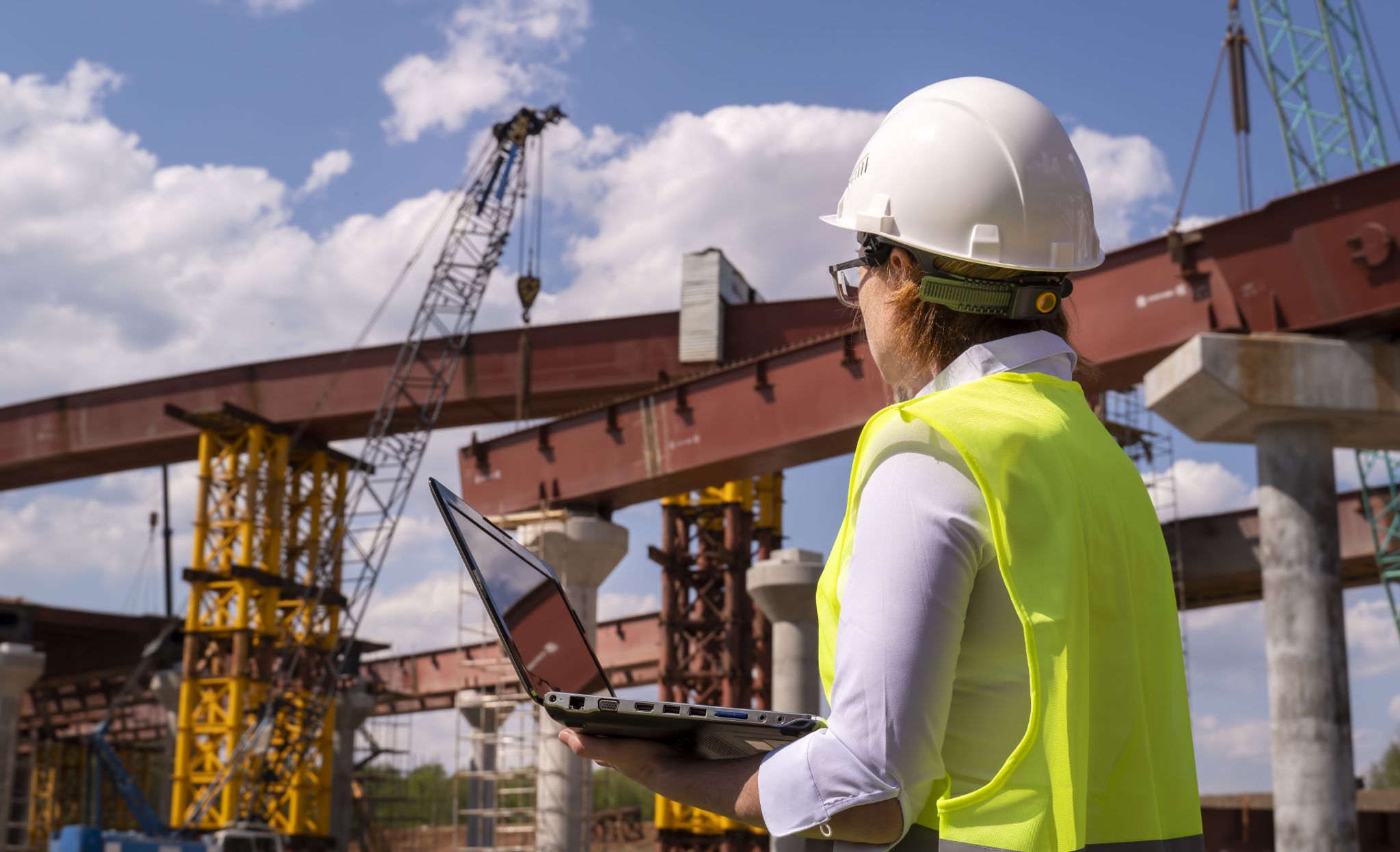A professional in a hard hat using a laptop at a bridge construction site